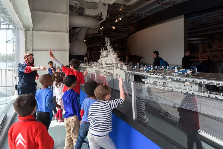 A group of kids are looking at the LEGO model of the Intrepid with a Museum educator who's wearing a pilot's helmet.
