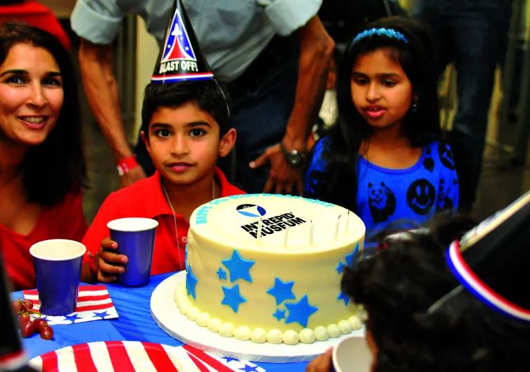 A kid wearing a party hat is seating with their family in front of a large cake with birthday candles on it.